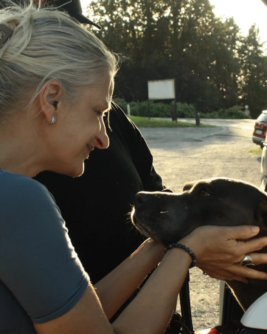 Sur un parking au retour de séance, l'éducatrice sourit en prenant doucement entre ses mains la tête d'un chien gris installé dans le coffre d'une voiture.